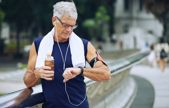 A healthy looking elderly man in workout costume standing on a blurred background of a park, holding a water bottle in one hand looks at the health activity tracker app on his other hand.